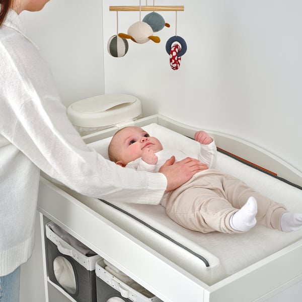 Caregiver with baby on SMÅGÖRA changing table, mobile above.