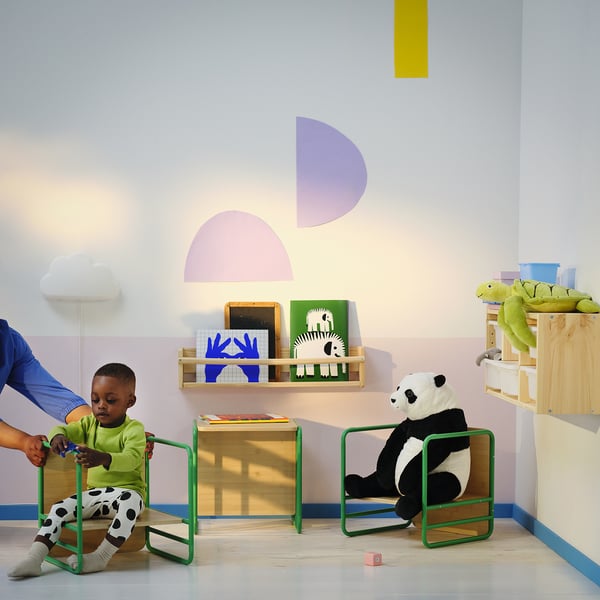 Playroom with child and panda teddy. Bright colours, shapes on wall. Child sits on green SMÅSKRAKE, adult holds childs hand.