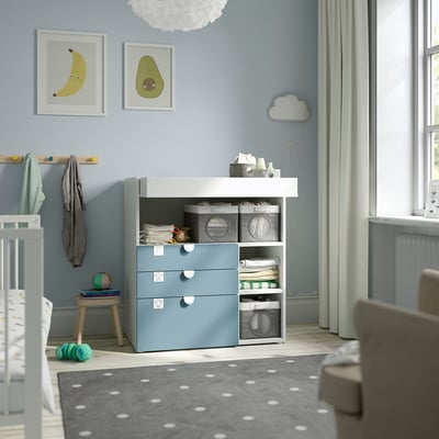 Blue SMÅSTAD changing table with storage, near window, next to crib and rug, child-friendly.