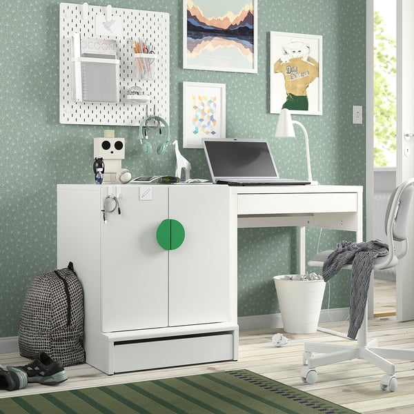 Small white SMÅSTAD cabinet, colourful knobs, holds kids items. Bright green wallpaper, artwork, laptop, desk. Cosy, organised space.
