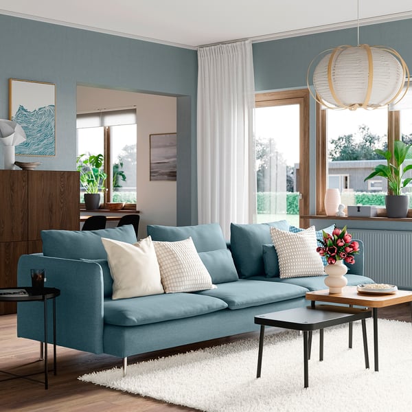 Modern living room with SÖDERHAMN blue sofa, pillows, white curtains, wooden cabinet, plant, and hanging lamp.