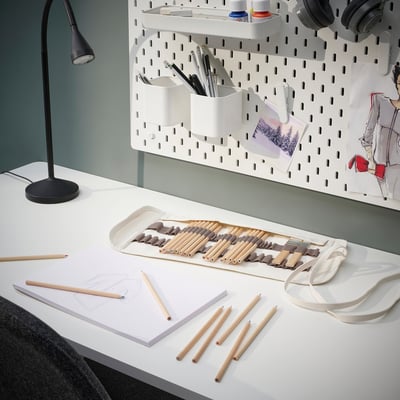 A white desk with SOLFÅGEL pencil roll, holding art supplies. Beside a white pegboard with stationery and sketches.