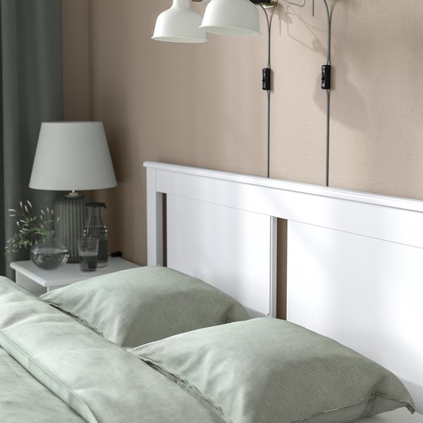 Minimalist bed frame with soft edges and high legs, shown with light green bedding. White table lamp and plant on bedside table, two wall lamps above.