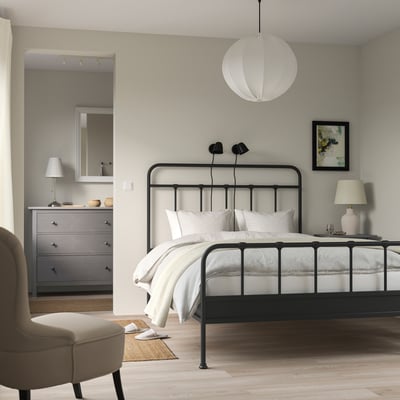 Modern bedroom with black curved bed, white linens, pendant lamp, grey walls, dresser, and armchair.