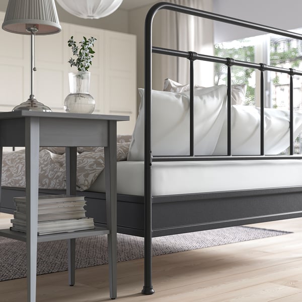 Black metal bed frame with curved headboard and footboard, white bedding, grey bedside table with lamp and books.