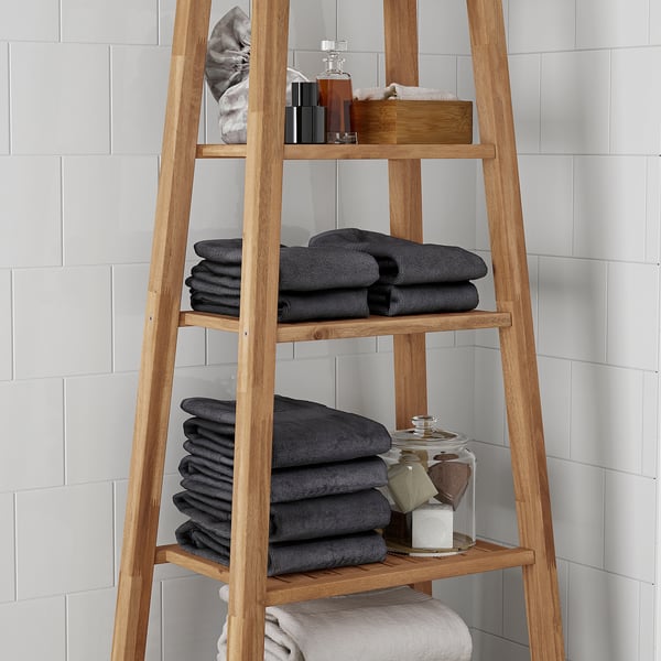 Wooden bathroom shelf unit with three tiers, holding folded dark towels, toiletries, and a glass jar, ideal for organising bathroom essentials.