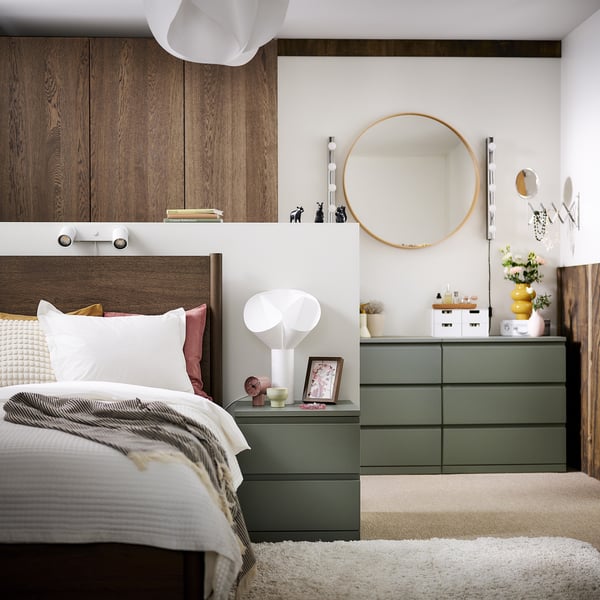 Modern bedroom with grey-green STORKLINTA bed, wooden headboard, pink pillows, and white bedding. Bedside table with lamp, mirror, rug, and storage.