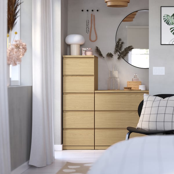 Modern bedroom with light wooden chest, hanging plants, mirror, lamp, and chequered pillows.