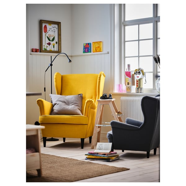 Yellow STRANDMON chair in cosy living room, large and inviting against grey chair and wooden ladder. Bookshelf by window adds colour.