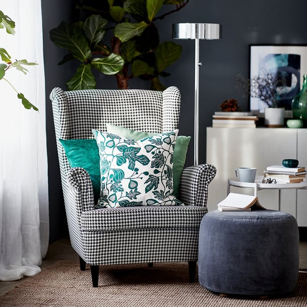 Modern living room with houndstooth STRANDMON chair, teal pillows, grey ottoman, floor lamp, and plant.