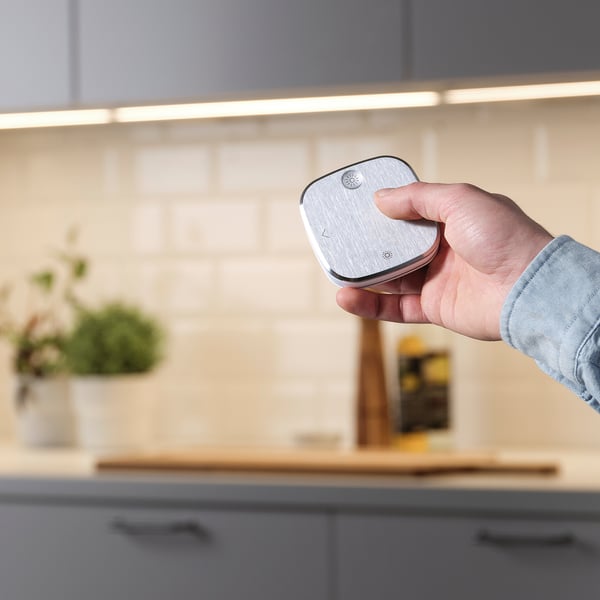 Hand holds STYRBAR remote, controlling smart kitchen lights, dimming and changing colour warmth.