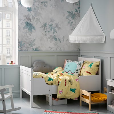 Childrens room with white SUNDVIK extendable bed, colourful bedding, stuffed cloud pillow, blue floral wallpaper, window, chair, and rug.