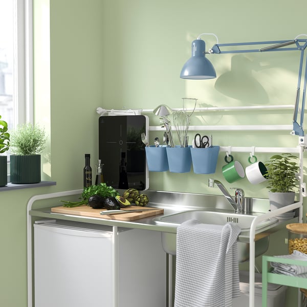 Small blue SUNNERSTA hanging organiser on kitchen wall, holding scissors, spoons, and mugs. Green wall and white sink.