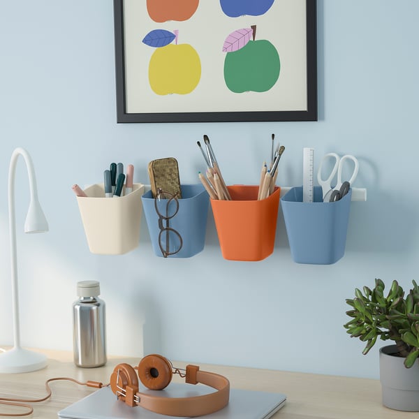 Four coloured SUNNERSTA wall containers for office supplies. Desk with lamp, headphones, plant. Apple picture above.