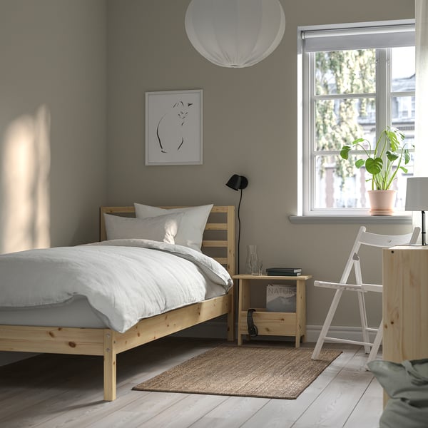 Minimalist grey bedroom with wooden TARVA bed, white linens, black art, small bedside table with lamp, and a potted plant on a desk.