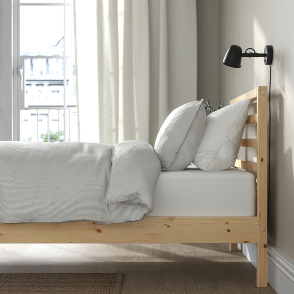 Bed by window, light-colored wood frame, white bedding, wall-mounted black lamp.