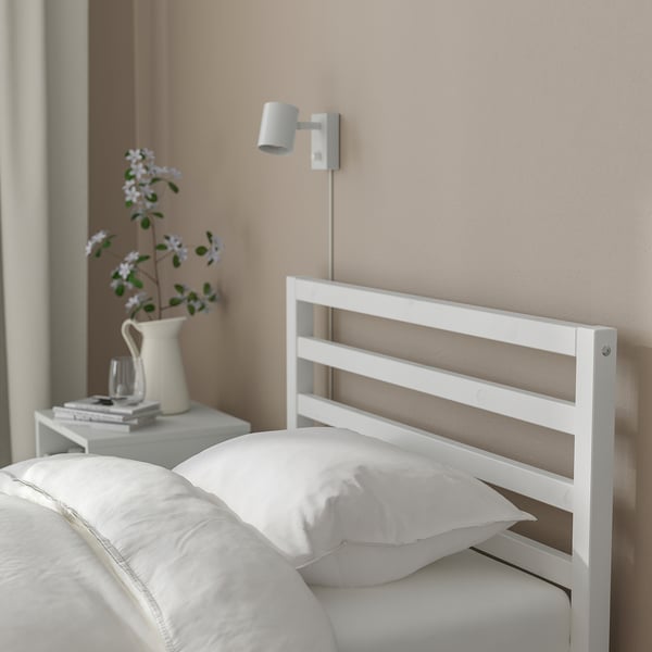 Minimalist white bed frame with tall headboard, wall light, white bedding, vase of flowers, and stack of books.