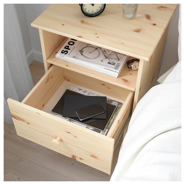 Wooden bedside table with open drawer showing a laptop, phone, and notebook. Top shelf has a magazine, watch, and glass.