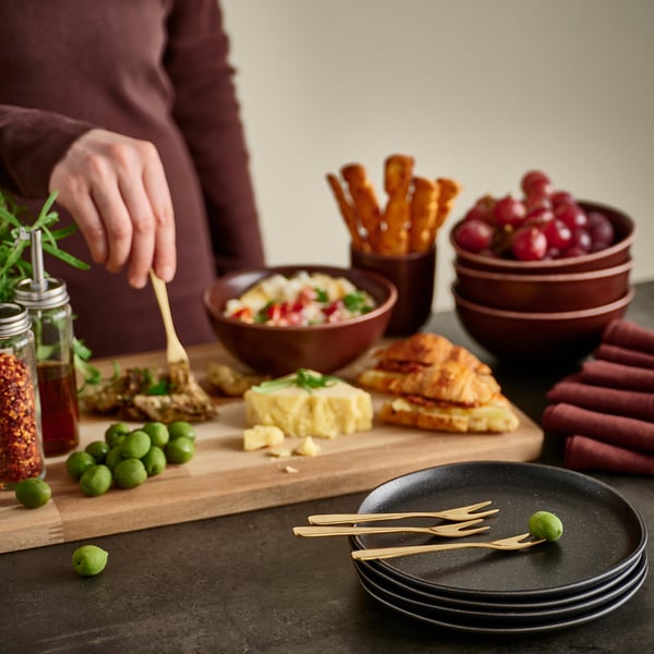 Person arranging food on a wooden board with TILLAGD brass-colored utensils and bowls.