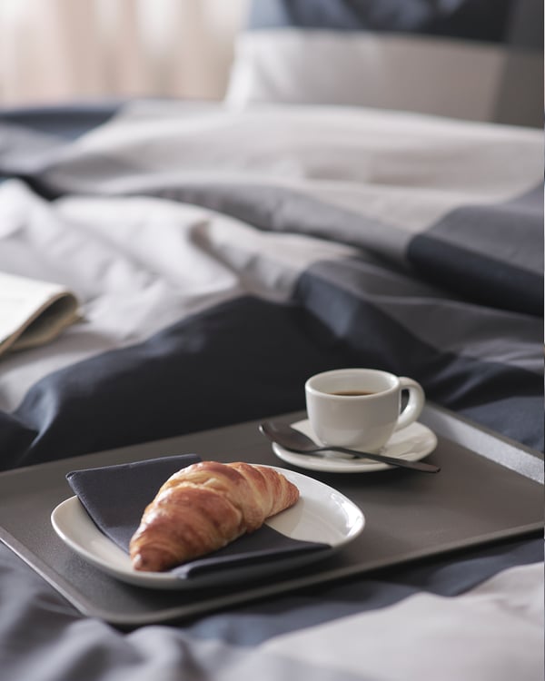 A black polypropylene plastic TILLGÅNG tray holding a pastry and a coffee cup on a bed.