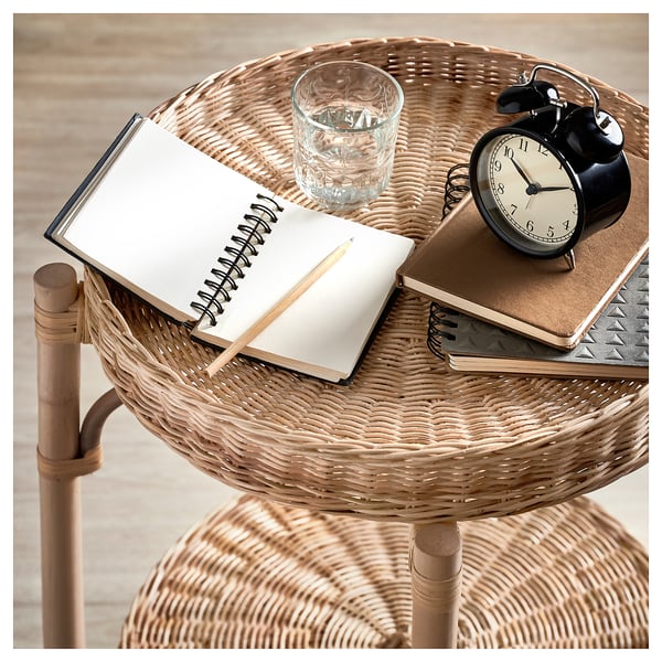 A TOLKNING natural rattan side table with a round wicker basket tray holding a notepad, glass of water, black alarm clock, and other items.