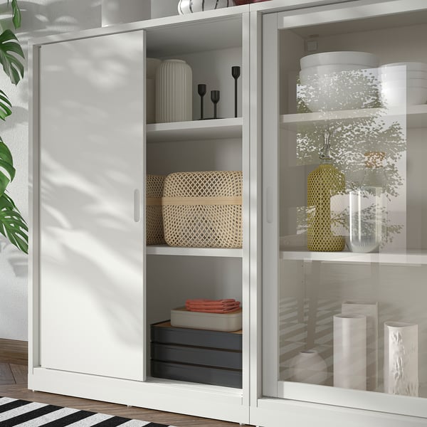 White TONSTAD storage unit with sliding doors and glass doors, featuring woven baskets, vases, and boxes. Modern, off-white design with clean lines.