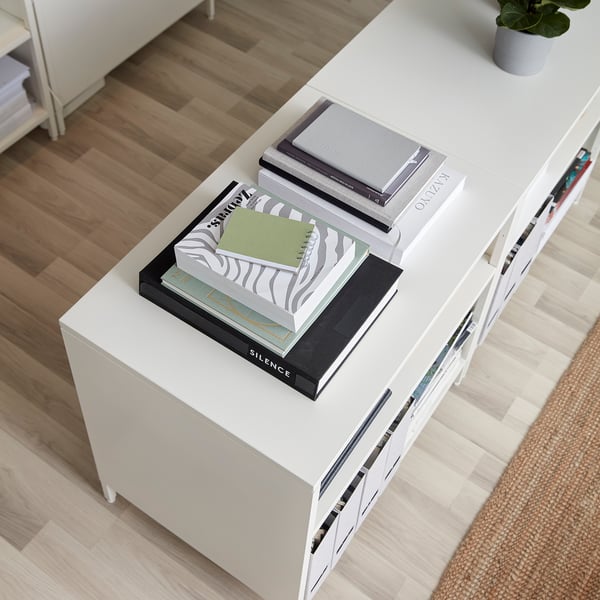 White TROTTEN cabinet with adjustable shelves, sliding doors & lock, housing books and a plant.