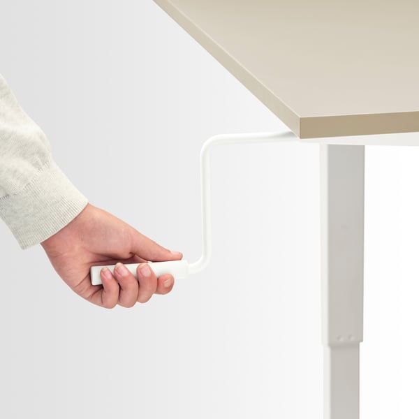 A hand adjusts a crank handle on a height-adjustable desk, allowing the user to switch between sitting and standing positions smoothly.