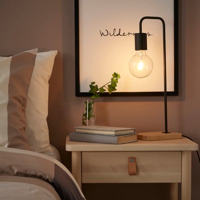 Cosy bedroom corner with black TVÄRHAND lamp on wood table, surrounded by books, plants, and pillows.