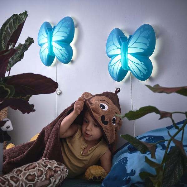 Kids hiding under towel, peeking between blue UPPLYST lights, surrounded by green plants and toys.
