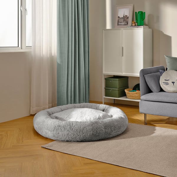 Cosy grey dog bed on wooden floor near window, part of living room with armchair and storage unit.