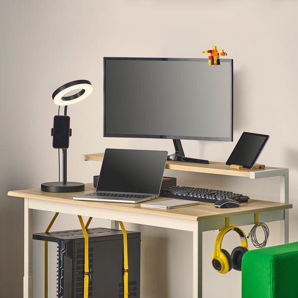 A compact gaming desk with adjustable height, a shelf for monitors, and cable management. Headphones hang on the side.