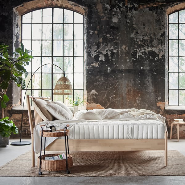 Relaxing bedroom with VÅGSTRANDA bed, white, wooden, plush mattress, woven rug, large arched windows, industrial wall, lamp, and plants.