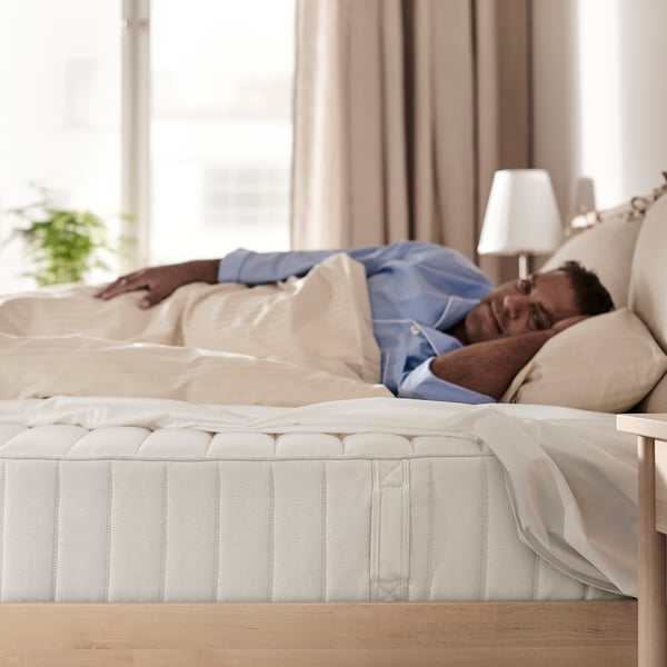 Person sleeping on a white mattress with beige pillow in a well-lit room with a window and a lamp.