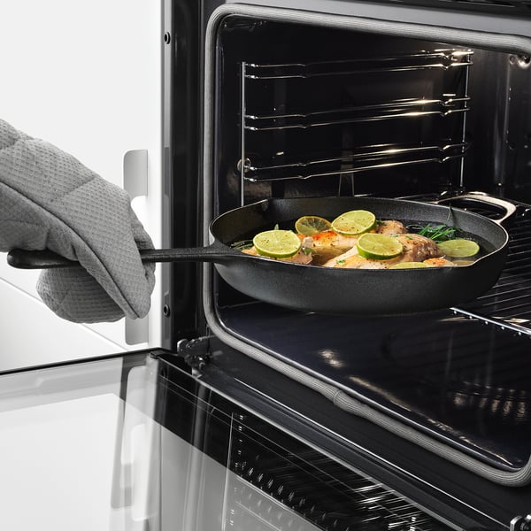 A gloved hand using oven mitts places a cast iron skillet into an oven. The skillet contains food and lime slices.