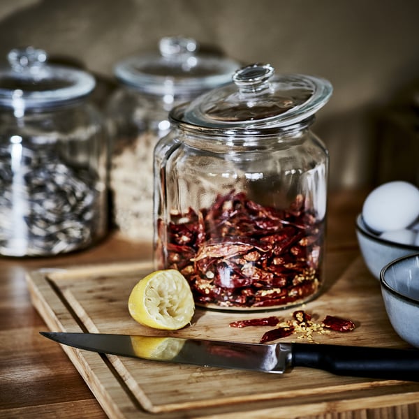 Clear glass jar with lid holds spices. Wooden chopping board has knife, lemon, and dried chilli flakes.