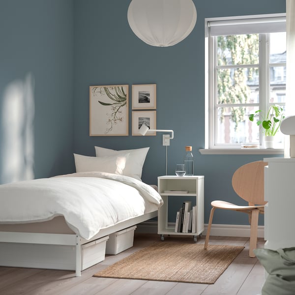 Teal bedroom with white bed, rug, jute mat, chair, bedside table, frames, lamp, window blinds.