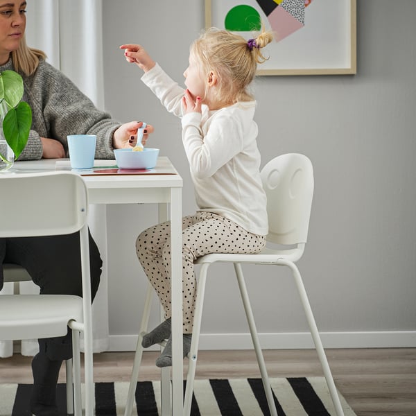 Child sitting on VILDGÅS junior chair, eating from a blue bowl. Mum sits beside, chair stacked under table.