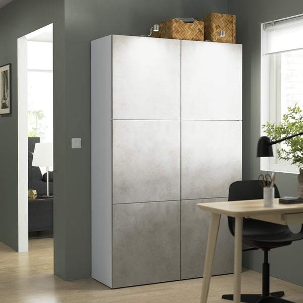Modern tall white BESTÅ storage unit in office. Sits between door and desk, higher than chair, with woven baskets on top.