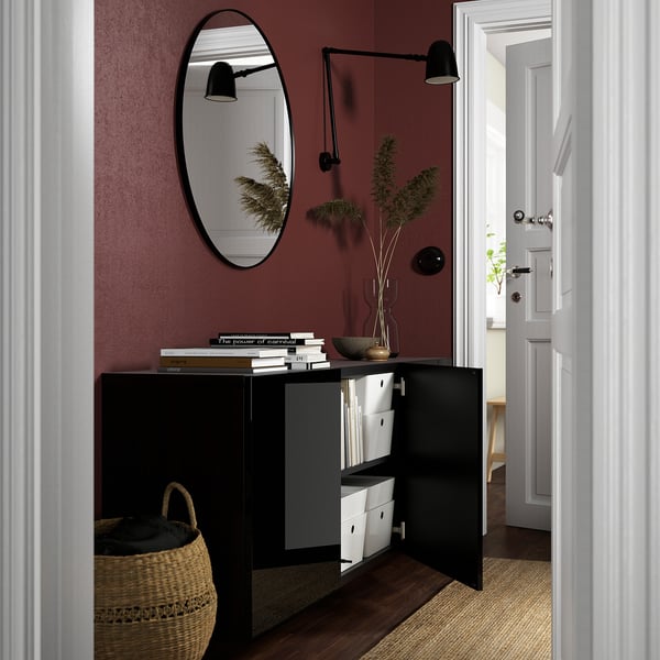 Black BESTÅ cabinet with open doors revealing adjustable shelves. A mirror, light, plant, and books sit on top.