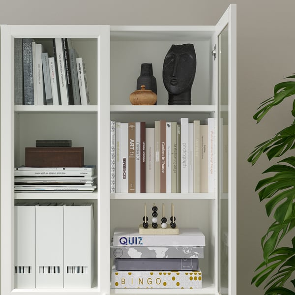 White BILLY bookcase with glass doors, displaying books, decorative items, and games, anchored to wall.