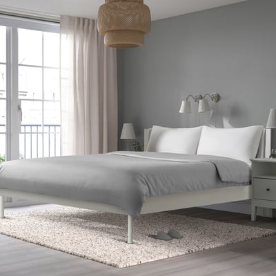 A modern bedroom with a white BRUKSVARA bed on a speckled rug. The bed has a cushioned headboard and ample space underneath for storage.