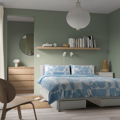 Modern bedroom with white bed, blue bedding, wooden dresser, and pendant light.
