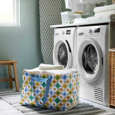 Large white and colourful patterned EKAÅN bag sits in laundry room, filled with towels, between washer and dryer. Folds flat, durable.