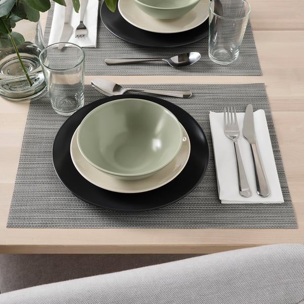 Two place settings with plain green bowls, white plates, glasses, and cutlery on woven placemats.