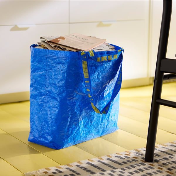 A blue ikea FRAKTA bag, filled with newspapers, placed on a patterned rug and next to a black chair on a wooden floor.