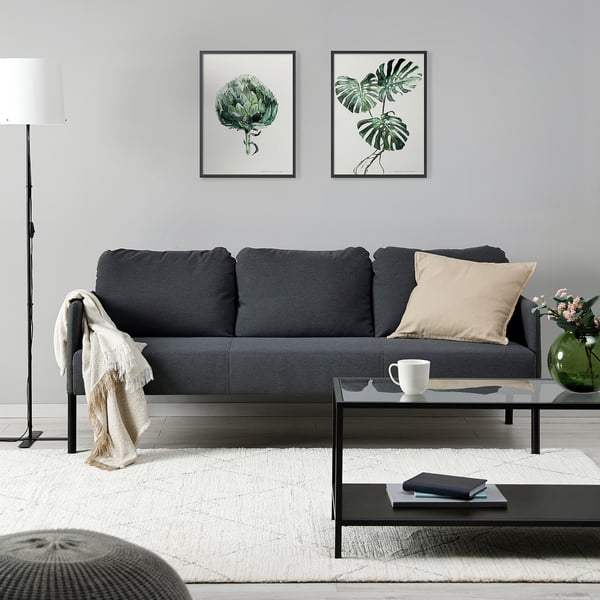 Modern living room: dark grey GLOSTAD sofa with beige cushions, white rug, glass coffee table, black lamp, plant art, cream throw, mug, and book.