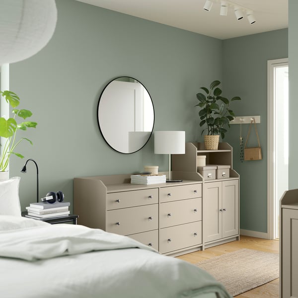 Bedroom with green walls HAUGA dresser and drawer combo in beige, topped with a lamp and plants. Round mirror above, white bed linens below.