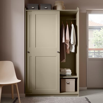 Beige HAUGA wardrobe with sliding doors, open showing clothes, boxes, and baskets. Light pink wall, white chair, modern minimalist room.