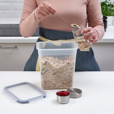 A person scoops granola from a large, clear plastic container on a kitchen counter, next to a small container of raspberries.